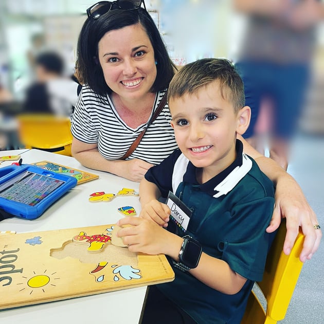 Photo of Darcy and his mum at school. darcy is at a table completing a puzzle and Chantal is behind him. 