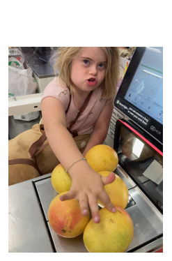 Photo of Eva at the self serve checkout at the supermarket, her hand is over the 5 mangos on the machine