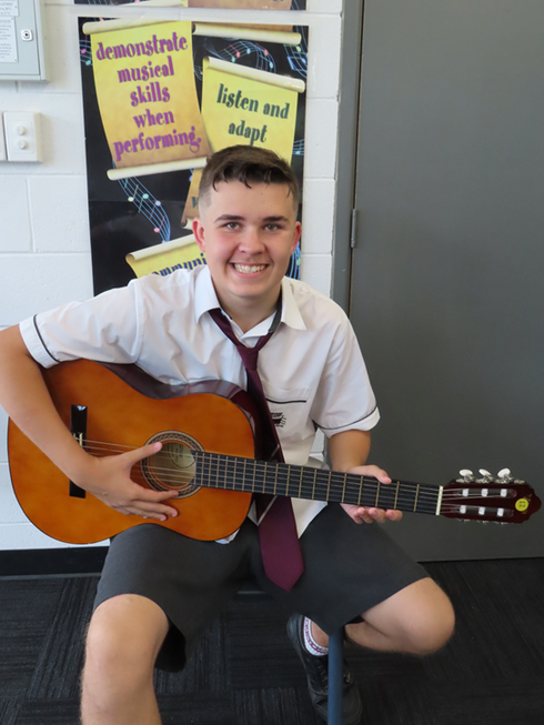 Photo of Sam sitting on a stool with his guitar in his school uniform. 